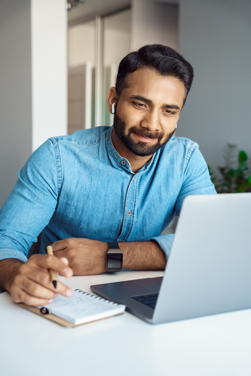 Portrait of young indian man student watching online webinar using laptop and earphone making noes in notebook. Video conference call, business training tutorial, coaching course concept